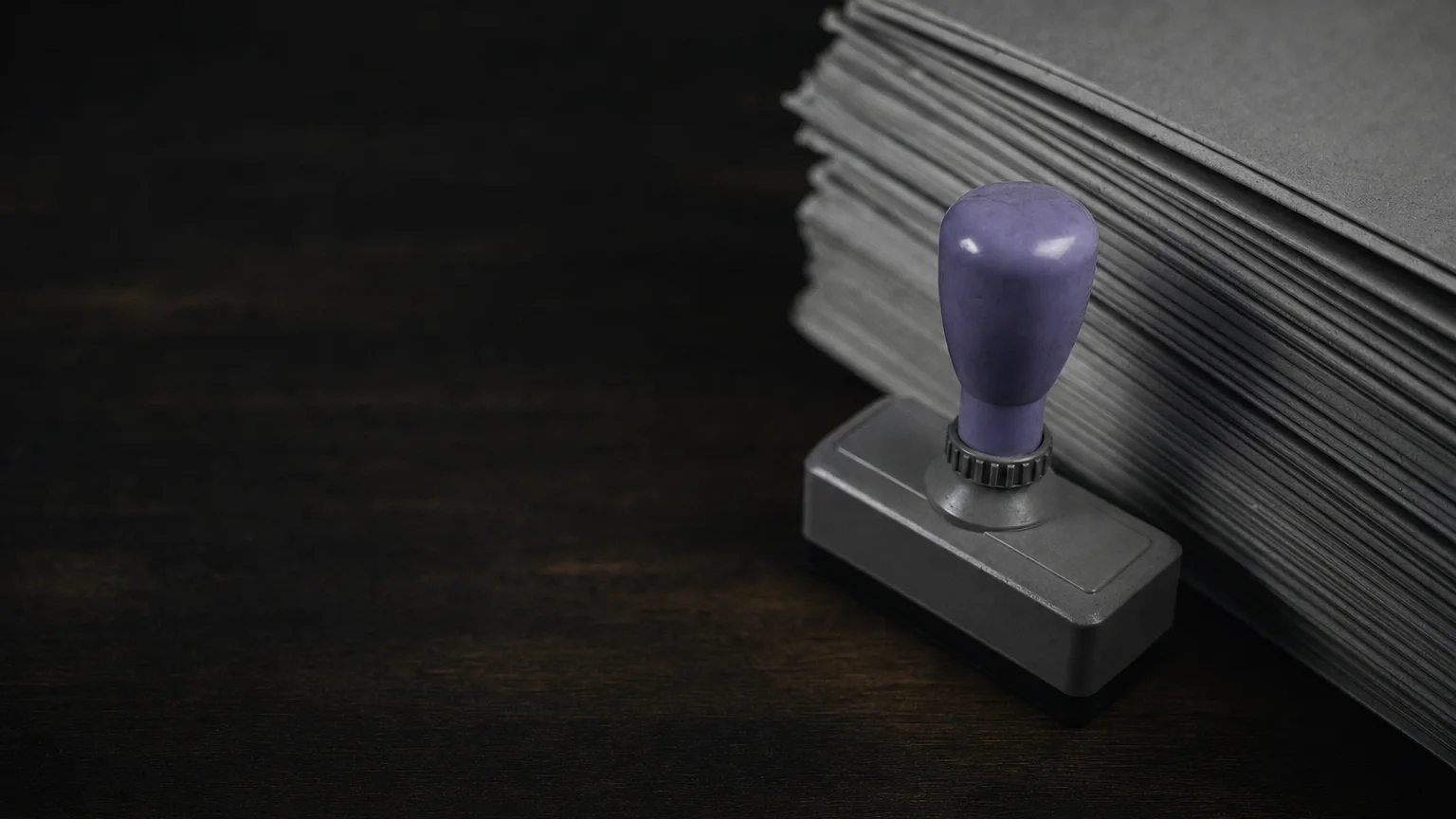 Photo of a rubber stamp with a violet handle next to a stack of grey forms on a dark wooden desk